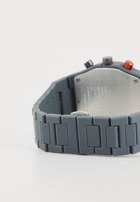 Orologio in silicone grigio con un cinturino testurizzato caratterizzato da anelli interconnessi. Retro metallico e hardware minimalista con tre pulsanti colorati.