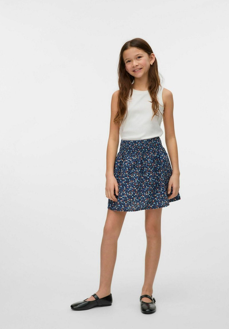 Top sin mangas blanco combinado con una falda de patrón floral azul. La falda tiene una cintura fruncida y cae a mitad del muslo. Zapatos negros completan el conjunto.