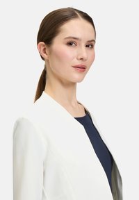 Witte blazer met een elegant ontwerp en sjalkraag, gecombineerd met een marineblauw topje eronder. De stof lijkt glad met minimale details.
