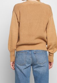 Beiger Strickpullover mit strukturiertem Muster, überschnittenen Schultern und gerippten Bündchen. Kombiniert mit hoch taillierten blauen Jeans, die zwei Gesäßtaschen haben.