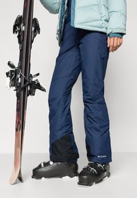 Tengerészkék sínadrág fekete akcentusokkal, vízálló anyagból készült. Egy személy áll egy síléccel és fekete sícipőkkel, szilárd csatokkal.