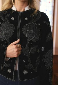 Juodas aksominis švarkas su gėlėmis siuvinėtomis detalėmis, karoliukais ir sidabro akcentais. Dizainas apima sudėtingus raštus ir trumpą ilgį.