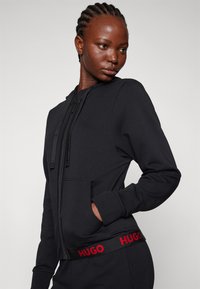Zwarte rits hoodie met voorpockets, trekkoord capuchon en een rode logoband op de taille. Heeft een zachte stof en een strakke pasvorm.