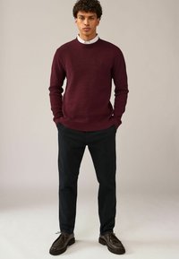 Burgundy kötött pulóver, kerek nyakkivágással, bordás mandzsettával, sötét nadrággal és barna fűzős cipővel párosítva; egy kis hímzett logóval rendelkezik.