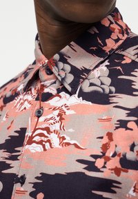 Πουκάμισο με γιακά που διαθέτει ένα λουλουδάτο μοτίβο σε κόκκινο, ροζ και γκρι πάνω σε σκουρόχρωμο ναυτικό φόντο. Η υφή του υφάσματος φαίνεται απαλή και υφαντή.