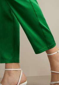 Pantalon en satin vert avec une texture lisse et une coupe ample, associé à des sandales blanches à lanières avec une boucle.