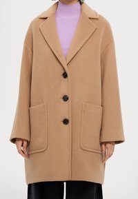 Abrigo de mezcla de lana beige con cuello de muesca, cuatro botones negros y bolsillos de parche en la parte frontal. Presenta una silueta relajada y oversize y mangas largas.