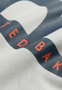 Baumwollstoff in Hellgrau mit einem marineblauen Block, auf dem "UNITED" und "BAKER" in fettem Orange gedruckt sind. Glatte Textur mit leichtem Glanz.