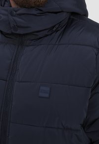 Marineblå polstret jakke med høj krave, der har lynlås og knapdetaljer. Inkluderer et Urban Classics logo-mærke på brystet.