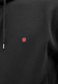 Felpa nera realizzata in tessuto morbido con un piccolo cerchio ricamato di rosso sul petto sinistro. Presenta un cappuccio con coulisse e polsini a coste.