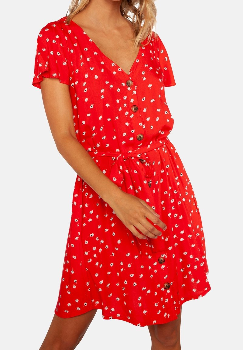 Robe fleurie rouge à manches courtes, col en V, avec détail de boutons, ceinture à nouer et petits motifs de fleurs blanches sur un tissu léger.