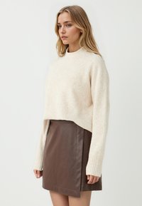 Maglione crema corto con polsini a coste abbinato a una minigonna in pelle marrone con spacco laterale. Texture morbida, design semplice.