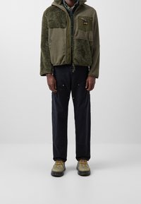 Zielona polarowa kurtka z teksturowaną, puszystą lewą stroną, kieszenią na piersi na zamek, w połączeniu z ciemnymi spodniami i jasnymi butami z żółtymi sznurowadłami.