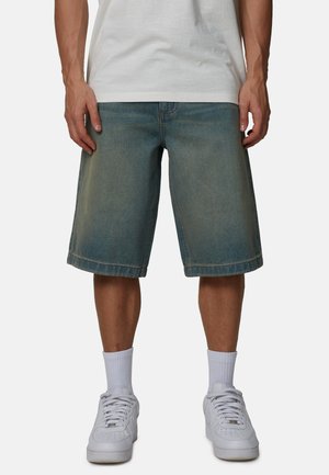 Mężczyzna ubrany w wyblakłe niebieskie szorty dżinsowe, białe skarpetki do kostek, białe trampki oraz białą koszulkę, stojący na jednolitym tle.