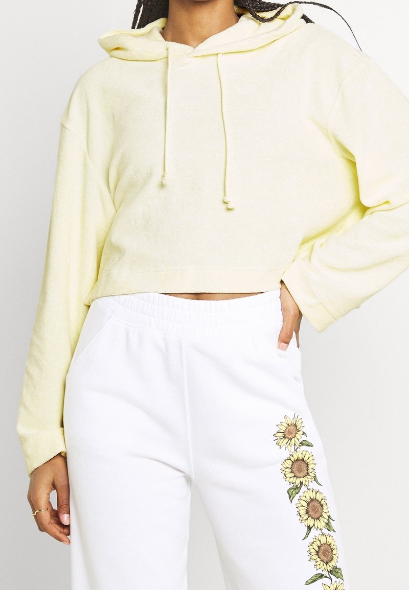 Sudadera amarilla corta con capucha ajustable, combinada con pantalones de chándal blancos que presentan gráficos de girasoles a lo largo de la pierna. Textura suave.
