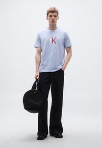 Ανοιχτό μπλε βαμβακερό t-shirt με κυκλικό λογότυπο "CK" σε κόκκινο, συνδυασμένο με μαύρο τζιν καμπάνα και στρογγυλή μαύρη τσάντα ώμου. Μαύρα παπούτσια.