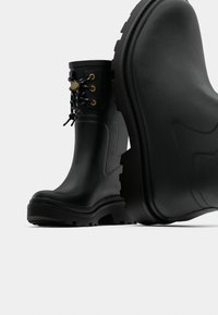 Μαύρα λαστιχένια μποτάκια αστραγάλου με υφή, που διαθέτουν χρυσούς μεταλλικούς τρύπες και αντίθετα μαύρα κορδόνια, με παχύ πέλμα για καλύτερη πρόσφυση.