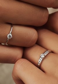 Deux bagues en argent sur des doigts, l'une avec un seul diamant rond et l'autre mettant en valeur plusieurs petites pierres le long de l'anneau.