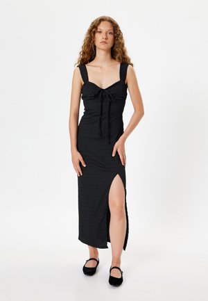 Vestido negro de tela texturizada, con silueta ajustada, escote cuadrado y abertura lateral. Combinado con zapatos negros.