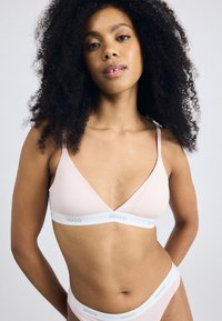 Lys rosa bralette med trekantet design og tynne stropper, med en hvit elastisk stropp med "HUGO"-logo. Glatt tekstur og minimalistisk stil.