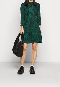 Grön leopardmönstrad klänning med långa ärmar och flera lager. Matchas med svart väska och kraftiga svarta skor med strukturerad sula.