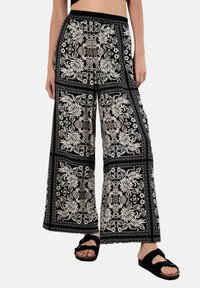 Schwarze weite Hosen mit einem kontrastierenden weißen Blumenmuster, elastischem Bund und weichem Stoff. Kombiniert mit schwarzen Sandalen.