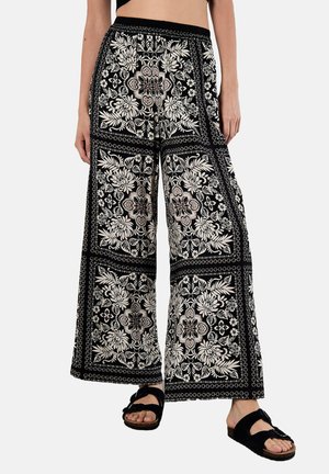 Schwarze weite Hosen mit einem kontrastierenden weißen Blumenmuster, elastischem Bund und weichem Stoff. Kombiniert mit schwarzen Sandalen.