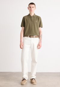 Olivenfarvet polo skjorte med tre knapper, hvide denim bukser, brun bælte og beige sko med snørebånd, stående på en lys grå overflade.