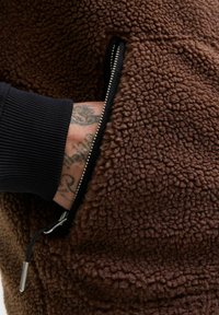 Hand mit Tattoos, teilweise in der Reißverschluss-Tasche einer braunen, strukturierten Fleecejacke, mit sichtbarem schwarzem Rippbündchen.
