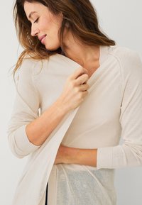 Cardigan leggero beige con scollatura a V e maniche lunghe. Presenta una consistenza morbida e una vestibilità comoda, indossato sopra una maglietta leggera.