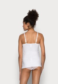 White camisole featuring adjustable thin straps, smooth texture, and a scalloped hem. Worn with white lace-trimmed panties.