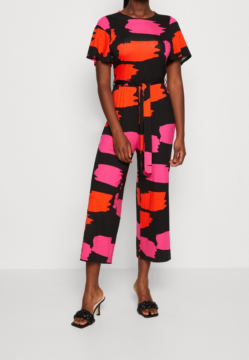 Femme portant une combinaison noire à manches courtes avec de larges motifs à coups de pinceau roses et rouges, ceinture nouée, jambes raccourcies, et sandales noires à talons hauts tressés.