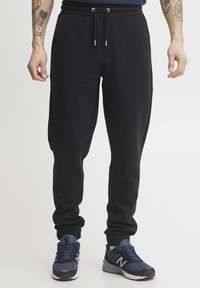 Zwarte joggingsbroek van zachte stof, met een trekkoord in de taille, gecuffed broekspijpen en zijzakken. Draagt met donkere sport schoenen.