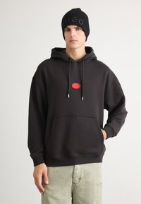 Felpa nera con una toppa con logo rosso, cappuccio con coulisse e tasca a marsupio; abbinata a pantaloni leggeri verde chiaro, texture, e un beanie in maglia nera.