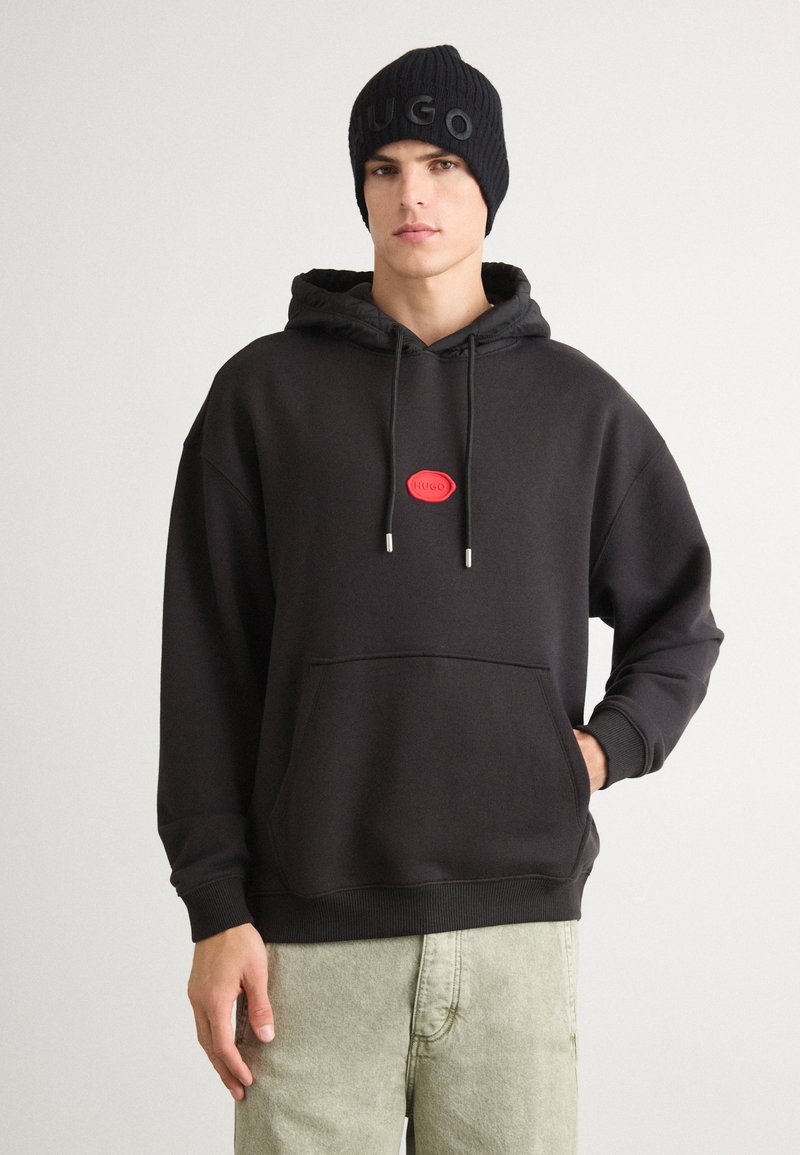 Felpa nera con una toppa con logo rosso, cappuccio con coulisse e tasca a marsupio; abbinata a pantaloni leggeri verde chiaro, texture, e un beanie in maglia nera.