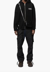 Sweat à capuche zippé noir avec le texte "PEQUS", pantalon cargo noir avec poches, et baskets marron. Comprend un sac bandoulière noir texturé.