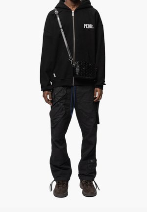 Sweat à capuche zippé noir avec le texte "PEQUS", pantalon cargo noir avec poches, et baskets marron. Comprend un sac bandoulière noir texturé.