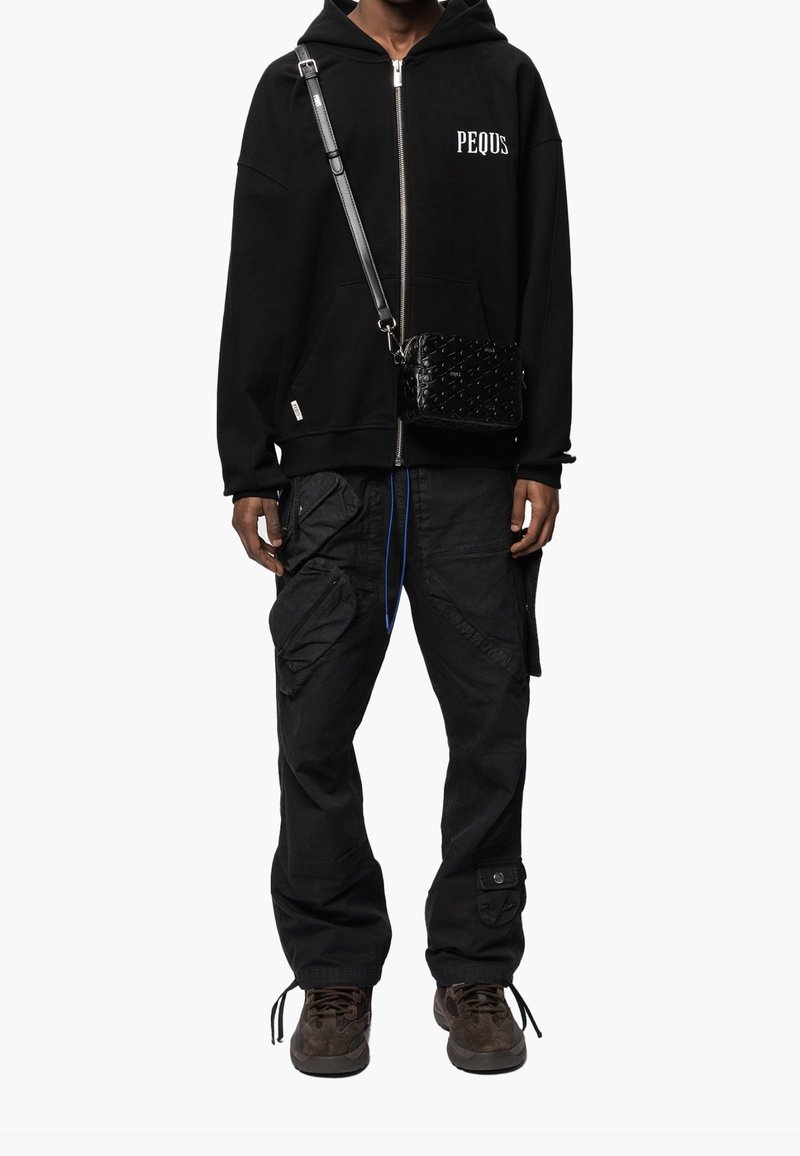 Sweat à capuche zippé noir avec le texte "PEQUS", pantalon cargo noir avec poches, et baskets marron. Comprend un sac bandoulière noir texturé.