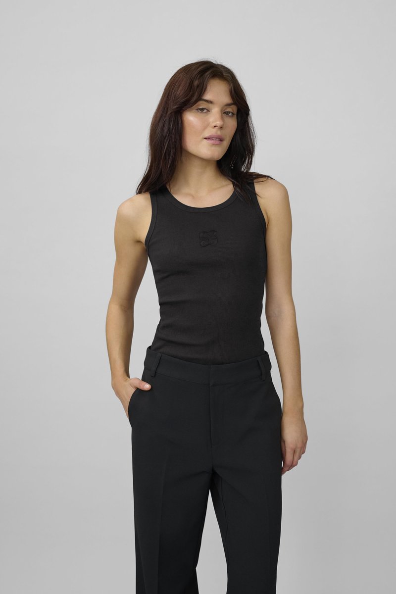 Junge Frau mit langen braunen Haaren, die ein eng anliegendes schwarzes ärmelloses Oberteil und hochtaillierte schwarze Hosen trägt, steht mit einer Hand in der Tasche.