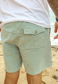 Shorts en velours côtelé vert clair avec une poche arrière et un détail de bouton, associés à un t-shirt blanc, debout sur une plage de sable.