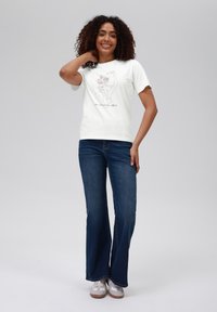 Hvit bomulls t-skjorte med et blomsterhåndgrafikk, kombinert med blå utsvingte jeans. Modellen smiler og poserer med en hånd på skulderen.