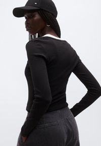 Musta pitkähihainen paita ribbineulosta, yhdistetty harmaisiin korkeavyötäröisiin housuihin. Mallilla on musta lippalakki ja hänellä on punotut hiukset.