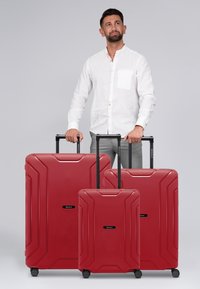 Set di tre valigie rigide di colore rosso, con superficie testurizzata, maniglie retrattili nere e quattro ruote per la mobilità.