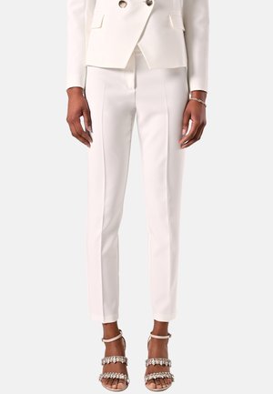 Personne portant un pantalon blanc sur mesure, un blazer blanc assorti et des talons ouverts avec des brides ornées de bijoux, debout devant un fond uni.