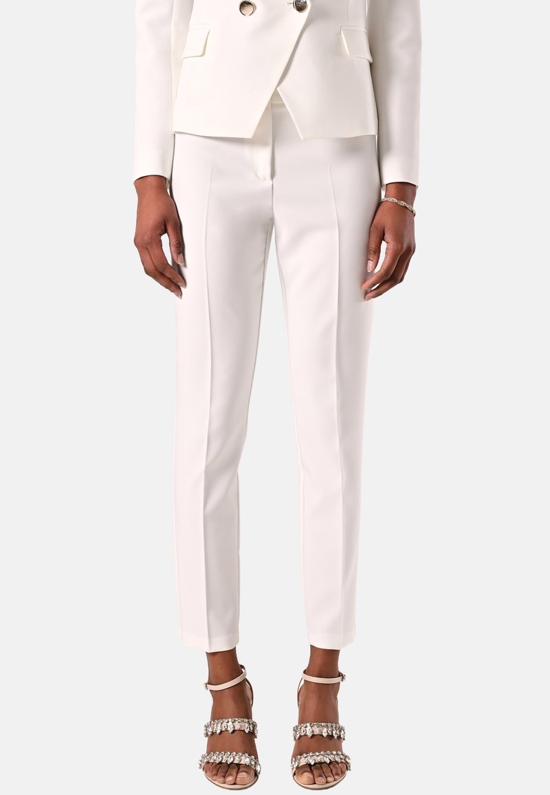 Personne portant un pantalon blanc sur mesure, un blazer blanc assorti et des talons ouverts avec des brides ornées de bijoux, debout devant un fond uni.