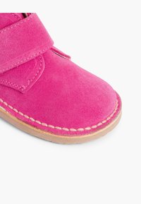 Pantof brunet din suede roz aprins, cu vârful rotunjit, având o curea lată din velcro și cusături bej de-a lungul marginii tălpii.