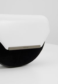 Petit sac bandoulière noir et blanc avec un fond en demi-cercle, un rabat angulaire et une plaque métallique de marque portant l'inscription "Ted Baker."