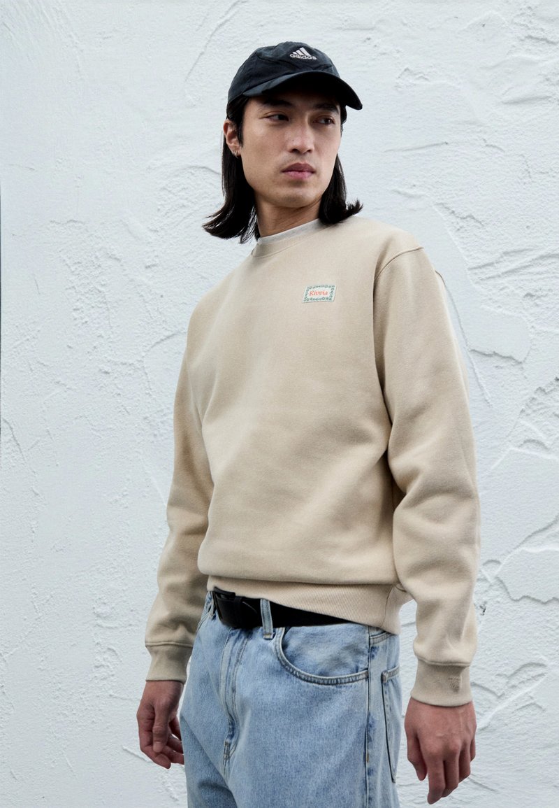 Sweat-shirt beige à col rond et poignets côtelés, avec un petit patch logo multicolore ; associé à un jean en denim bleu clair.
