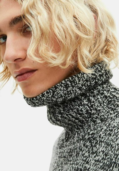 Artimo plano nuotrauka asmens su banguotais šviesiais plaukais, dėvinčio storą juodai baltą megztinį su aukštu kaklu, žvelgiančio šiek tiek šoną.