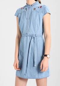 Denim dress with short sleeves, light blue color, embroidered details on the chest, and a belted waist. Button-down front, casual design.